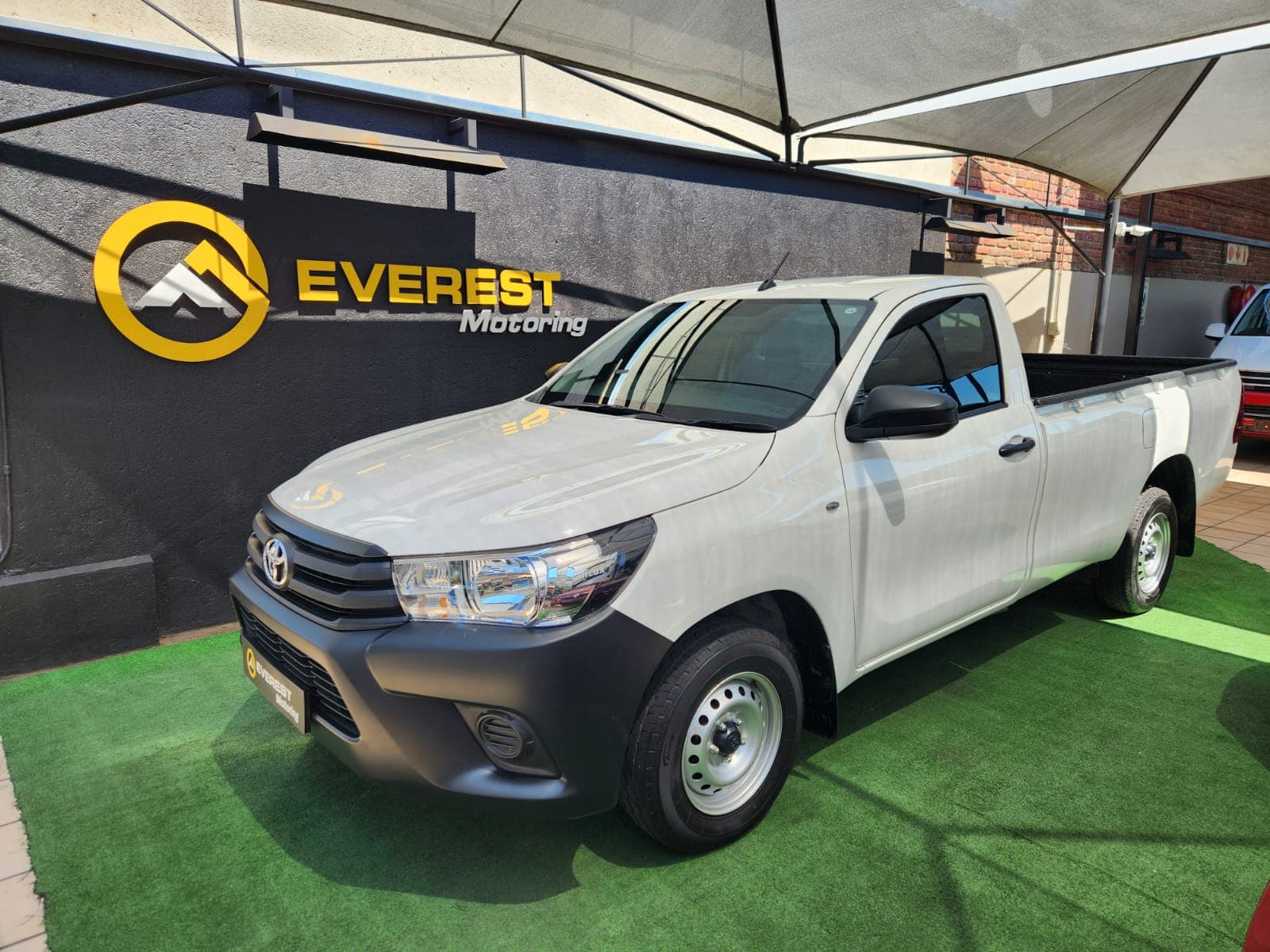 White 2025 Toyota Hilux 2.4 GD Single-Cab, front three-quarter view, parked on green turf at dealership.
